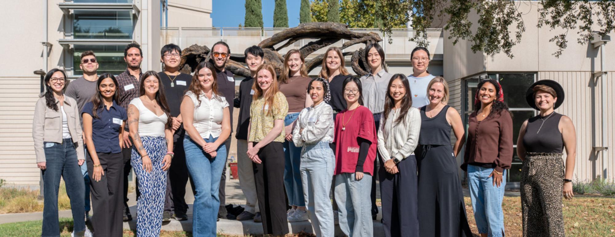 Outdoor group photo of about 25 adults standing on grass by a building and tree.